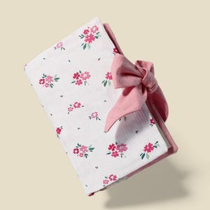 Pode incluir: Um cobertor de bebê branco dobrado com um padrão floral rosa e um grande laço rosa. O cobertor apresenta um padrão repetido de flores rosa e folhas verdes. O laço é feito de um material macio e texturizado.