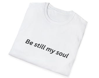 May include: White t-shirt with the text "Be still my soul" printed in black. The shirt is folded and appears to be made of a soft, lightweight material. The design is simple and minimalist.