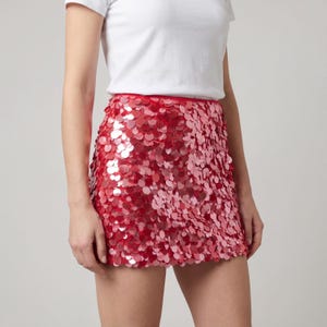 May include: A red sequin mini skirt. The skirt is covered in overlapping, shiny sequins in shades of red. The skirt is paired with a white t-shirt. The skirt has a scalloped hemline.