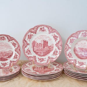 Könnte beinhalten: Ein Satz Vintage-Keramikteller mit einem rot-weißen Transferdruck. Die Teller haben einen gewellten Rand und eine Landschaftsszene. Das Set umfasst Essteller und kleinere Teller, die auf goldenen Tellern gestapelt sind.