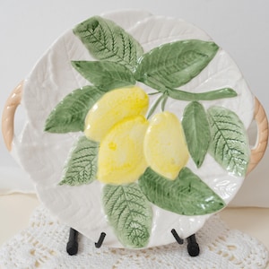 Könnte beinhalten: Weißer Keramik-Servierteller mit Zitronen- und Blattdesign. Der Teller hat ein geprägtes Blättermuster an den Rändern und zwei geflochtene Griffe. Die Zitronen sind gelb und die Blätter grün.