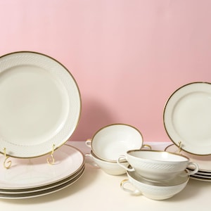 Pode incluir: Um conjunto de louça composto por pratos e taças na cor creme com um padrão de diamante texturizado nas bordas e acabamentos em tom dourado. A coleção inclui pratos grandes empilhados, pratos menores e taças de sopa com duas alças.