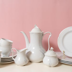 May include: A white porcelain tea set with a floral design, including a teapot, creamer, sugar bowl, cups, and plates. The plates are stacked on a gold-coloured stand. The set is displayed on a white surface against a pink background.