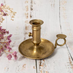 Può includere: Un portacandele vintage in ottone con base rotonda e manico. Il portacandele ha uno stelo cilindrico e un piccolo pomello. Fiori secchi con fiori rosa e bianchi sullo sfondo.