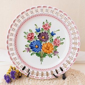 Könnte beinhalten: Ein weißer Keramikteller mit einem handgemalten Blumenstrauß in Rosa, Blau, Orange und Violett sowie grünen Blättern. Der Tellerrand ist mit einem gitterartigen Muster, rosa Akzenten und einem gewellten Rand gestaltet.