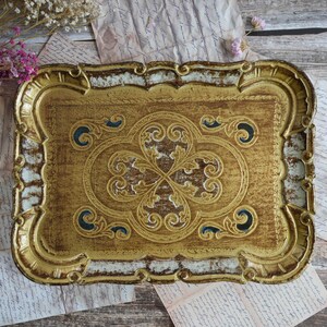 Pode incluir: Uma bandeja retangular com acabamento dourado envelhecido e uma borda ornamentada em relevo. O centro apresenta um padrão de volutas florais simétricas em relevo com detalhes em azul escuro. As bordas têm um design festonado com toques brancos.