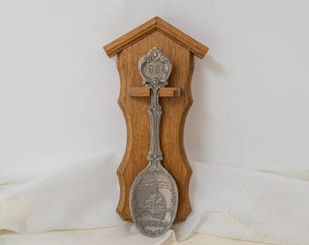 May include: A silver-toned metal souvenir spoon featuring an embossed building and the year 1984, mounted on a shaped light brown wooden wall plaque with a peaked roof design. The spoon handle has ornate detailing and the year 1984 at the top.