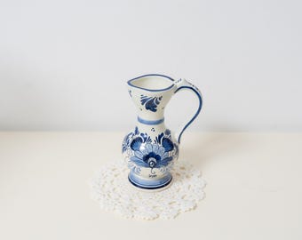 May include: A small ceramic pitcher featuring a white base with intricate blue floral patterns. The vessel has a rounded body, a flared rim, and a curved handle. The hand-painted design includes stylized petals and leaves around the base and neck.