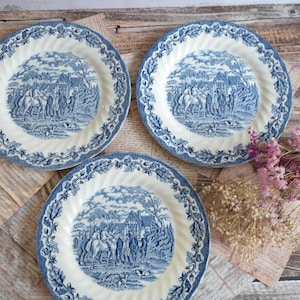 Può includere: Tre piatti rotondi in ceramica con un motivo a trasferimento blu e bianco. Ogni piatto mostra una scena dettagliata di figure a cavallo vicino a un edificio, circondata da un bordo floreale decorativo con un design a bordo smerlato e scanalato.