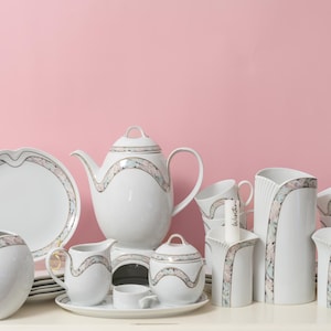 Könnte beinhalten: Ein weißes Porzellan-Teeservice mit einem dekorativen Design in Rosa, Grau und Gold. Das Set umfasst eine Teekanne, Tassen, eine Zuckerdose, einen Sahnekännchen, einen Servierteller und eine Vase. Das Set wird auf einer weißen Oberfläche vor einem rosa Hintergrund präsentiert.