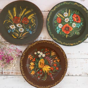 Puede incluir: Tres platos decorativos pintados a mano con diseños florales. Uno es verde oscuro con amapolas rojas, margaritas blancas y trigo amarillo. Otro es marrón con flores amarillas y naranjas. El tercero es verde oscuro con un borde floral.