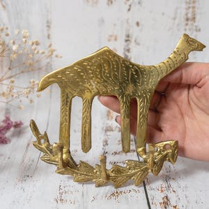 May include: A gold-toned metal wall hook shaped like a camel with an etched texture on its body. The base of the hook features a decorative leaf-like design with three individual prongs extending upward for hanging items on a wall surface.