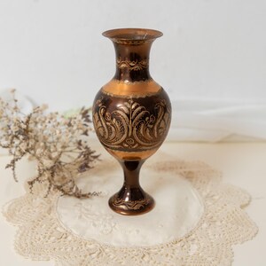 Könnte beinhalten: Eine dunkelbraune Metallvase mit ausgestelltem Rand und einem Standfuß. Die Vase hat ein poliertes, kupferfarbenes Band um die Mitte und den Hals, das mit filigranen, geätzten goldfarbenen Blumen- und Rankenmustern auf dem Körper verziert ist.