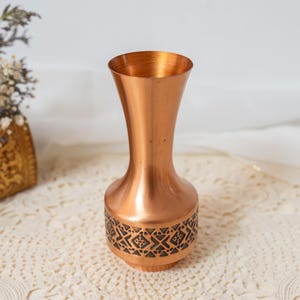 Könnte beinhalten: Eine Vase aus kupferfarbenem Metall mit einem schlanken Hals und einem breiteren Fuß. Der untere Teil der Vase weist ein dunkles, komplexes geometrisches Muster auf, das in die Metalloberfläche eingraviert ist. Die Oberfläche ist glatt und glänzend.