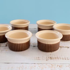 May include: A set of six round ceramic ramekins featuring a two-tone design with a cream-coloured interior and rim, and a dark brown exterior with vertical ribbed detailing. The small bowls are arranged in a group, showcasing their uniform shape.