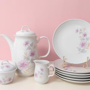Può includere: Un servizio da tè in porcellana bianca con un motivo floreale. Il set include una teiera, una zuccheriera, una lattiera e dei piatti. I piatti sono impilati su un supporto dorato. Il motivo floreale presenta fiori rosa e grigi.