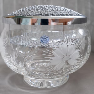 Op de afbeelding: Een heldere glazen bloemenvaas met een zilveren metalen rand en een gaas bovenkant. De vaas heeft een ronde vorm met geëtste bloemmotieven. Een blauw en wit label is zichtbaar aan de voorkant.