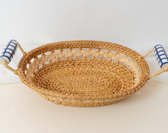 May include: An oval-shaped woven rattan basket featuring a natural light brown finish. The basket includes two side handles made of white ceramic with a blue grid pattern, attached to the rim with woven rattan loops and a decorative braided border.