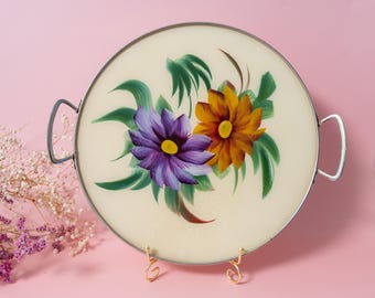 May include: A round serving tray with a cream-colored surface featuring a hand-painted design of one purple flower and one orange flower with green leaves. The tray has a thin silver-toned metal rim and two silver-toned metal side handles.