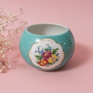 Może przedstawiać: Okrągła, turkusowa ceramiczna miska z kwiatowym wzorem. Miska posiada białą, falistą kartuszę z bukietem czerwonych, fioletowych i żółtych kwiatów. Obrzeże jest wykończone złotem. Tło jest w kolorze jasnoróżowym.