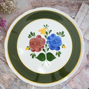 Op de afbeelding: Een ronde keramische bord met een wit middenstuk voorzien van een handgeschilderd bloemmotief van een bruine roos, een blauwe roos en een gele klokvormige bloem met groene bladeren. Het bord heeft een brede donkergroene rand met een gele lijn.