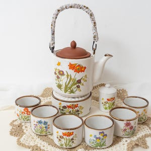 Può includere: Un servizio da tè vintage con una teiera bianca con coperchio e manico marrone, ornata da motivi floreali. Il set include tazze abbinate, uno scaldavivande e un piccolo contenitore, tutti decorati con fiori colorati. Il set è posto su un centrino decorativo.
