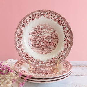 Può includere: Una pila di quattro piatti antichi con un motivo bianco e rosso. I piatti hanno un bordo smerlato con un motivo floreale. Il centro dei piatti presenta una scena pastorale rossa e bianca. I piatti sono esposti su una superficie di legno bianco.