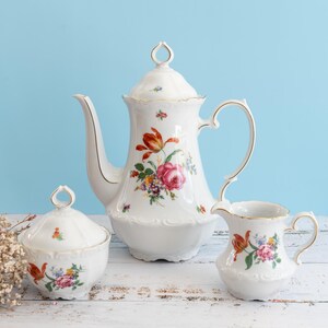 May include: A white porcelain tea set with floral designs, including a teapot, sugar bowl, and creamer. The set features red, pink, and yellow flowers with gold accents. The background is a light blue.