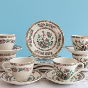 Può includere: Un servizio da tè con tazze e piattini in ceramica color bianco sporco, decorati con motivi floreali rosa, foglie verdi e bordi geometrici marroni. Una pila di piatti abbinati al centro presenta un motivo floreale centrale e una bordura.