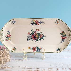 Puede incluir: Bandeja rectangular crema para servir con un diseño floral. La bandeja tiene un borde verde y está sostenida por un soporte de metal dorado. El diseño floral presenta flores rojas, moradas, amarillas y azules.