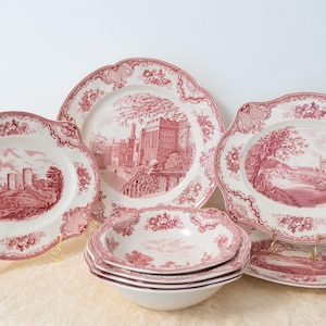 May include: A collection of antique white and red transferware dishes. The set includes plates and bowls with a scalloped edge and a detailed landscape scene. The dishes are arranged on a cream-coloured surface.