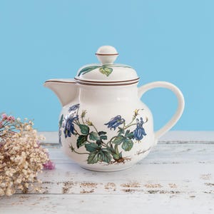 Può includere: Una teiera in ceramica bianca con un'illustrazione botanica di fiori di aquilegia blu con foglie e radici verdi. La teiera presenta un coperchio abbinato con un piccolo pomello, un manico curvo e un bordo marrone lungo la parte superiore.