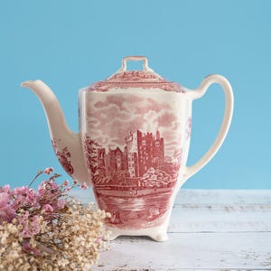 Può includere: Una teiera in ceramica color crema con una dettagliata stampa rossa che raffigura un paesaggio con castello. La teiera è dotata di coperchio coordinato, manico curvo, beccuccio lungo e poggia su piccoli piedini.