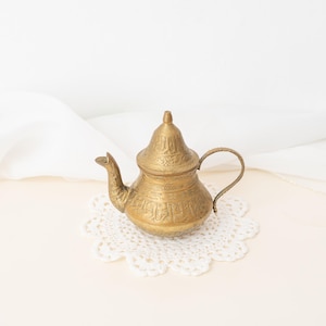 Peut inclure: Petite théière en laiton doré, ornée de motifs gravés et d'un couvercle en dôme. Elle repose sur un napperon en dentelle blanche. Le fond est de couleur neutre, avec un tissu blanc drapé. Objet décoratif pour la maison.