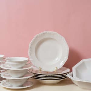 Peut inclure: Ensemble de vaisselle en porcelaine blanche, comprenant des soupières avec anses, des soucoupes, des assiettes et un saladier. Les pièces ont un bord décoratif festonné et sont disposées sur une surface blanche sur fond rose.