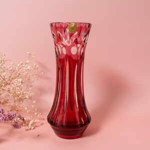 Può includere: Un vaso alto di vetro rosso rubino con la parte superiore svasata e un design strutturato e a coste. Il vaso ha una base rotonda e una forma sottile e allungata. Lo sfondo è rosa tenue, con rametti di fiori bianchi e viola a sinistra.