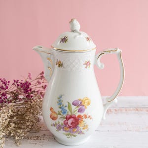 May include: A white porcelain coffee pot featuring a colorful floral bouquet pattern, gold trim on the handle, spout, and lid, and a small bird figurine perched on top of the lid. The body has a subtle embossed lattice texture near the rim.