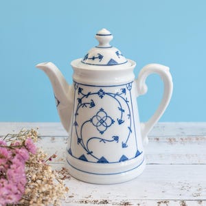 May include: A white porcelain teapot featuring a delicate blue floral and geometric pattern. The teapot has a matching lid with a small knob, a curved handle, and a tapered spout. The design includes intricate blue lines and small floral motifs.