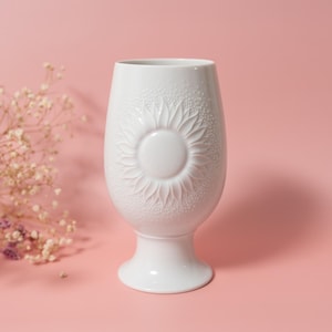May include: A white ceramic vase with a sunflower design. The vase has a wide, rounded body and a pedestal base. The sunflower is embossed on the front. The background is a solid pink color.