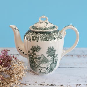 Op de afbeelding: Een crèmekleurige keramische theepot met een donkergroen transferpatroon van een landelijk landschap met bomen en figuren. De theepot heeft een bijpassend deksel met een knop, een gebogen handvat en een tuit, allemaal versierd met bloemmotieven.
