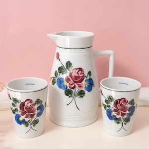 Op de afbeelding: Een witte keramische kan en twee bijpassende bekers, elk versierd met een handgeschilderd bloemmotief met een centrale rode roos omringd door kleinere blauwe bloemen en groene bladeren, afgewerkt met een dunne donkergroene rand.