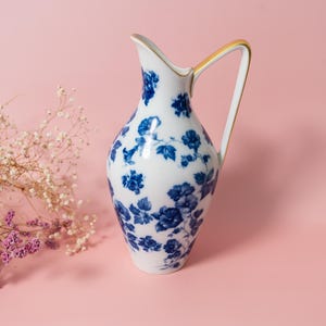 Op de afbeelding: Een witte porseleinen kan met een delicaat blauw bloemmotief over het oppervlak. De kan heeft een gebogen handvat met een goudkleurige rand langs de bovenkant en een taps toelopende tuit, wat zorgt voor een klassiek en glad ontwerp.