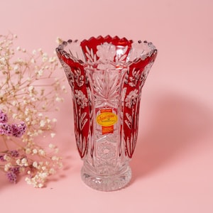 May include: A clear crystal vase with red detailing and floral patterns. The vase has a wide, scalloped rim and a round base. A sticker with text is visible on the front. The vase is displayed on a pink background.