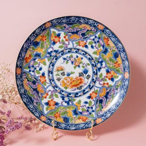 May include: A round ceramic decorative plate featuring a central floral motif surrounded by intricate patterns of orange, blue, and green flowers and leaves. The plate has a dark blue border with gold trim and rests on a gold metal display stand.