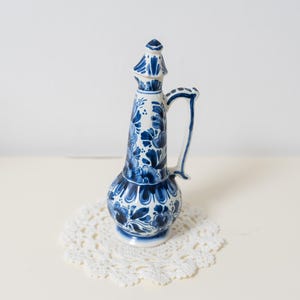 Może przedstawiać: Wysoki, niebiesko-biały ceramiczny dzbanek z uchwytem i ozdobną pokrywką. Dzbanek ma kwiatowy wzór i jest umieszczony na białej, okrągłej serwetce. Dzbanek ma około 25 cm wysokości.