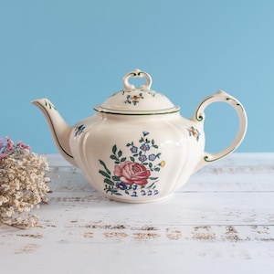 May include: Cream-colored ceramic teapot with a floral design featuring a red rose and blue flowers. The teapot has a curved spout and handle, and a matching lid. The background is a light blue, with dried flowers on the left.