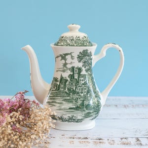 Puede incluir: Una tetera de cerámica blanca con un detallado patrón de paisaje en transferencia verde que muestra edificios y árboles. La tetera incluye una tapa a juego con diseño floral, un asa curva y un pico largo, exhibiendo un estilo clásico.