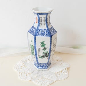 Op de afbeelding: Witte en blauwe keramische vaas met achthoekige vorm. De vaas heeft bloemen- en landschapsontwerpen, evenals Chinese karakters. Geplaatst op een witte, kanten onderzetter.