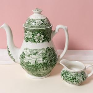 May include: A white ceramic teapot and creamer set with a green floral and landscape design. The teapot has a lid and a curved spout. The creamer has a small handle. The set is on a white surface with a pink background.