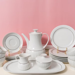 Op de afbeelding: Een wit porseleinen theeservies met een decoratieve zilveren rand. De set bevat een theepot, kopjes, schotels, borden, een melkkan, een suikerpot en een serveerschaal. De theeset is geplaatst op een wit oppervlak tegen een roze achtergrond.